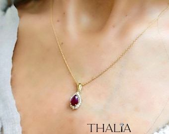 Op de afbeelding: Een gouden ketting met een peervormige robijn hanger omringd door een halo van kleinere diamanten. De hanger zit om de nek van een vrouw. De tekst "THALIA" is zichtbaar in de rechterbenedenhoek van de afbeelding.