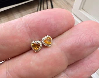 Puede incluir: Un par de pendientes de oro en forma de corazón con una pequeña piedra preciosa amarilla en el centro de cada corazón. Las piedras preciosas están rodeadas por un halo de piedras preciosas blancas más pequeñas.