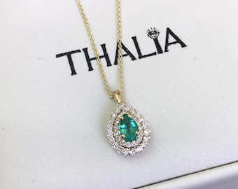 May include: A pear-shaped emerald gemstone pendant necklace with a double halo of diamonds set in yellow gold. The pendant is on a delicate chain and is displayed in a white box with the word "THALIA" printed on it.