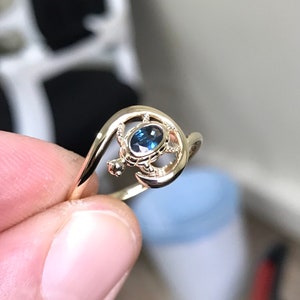 Peut inclure: Une bague dorée ornée d'un motif de tortue. La tortue est ornée d'une pierre précieuse ovale bleue. L'anneau de la bague s'enroule autour de la tortue. La bague est tenue par les doigts d'une personne. Le design est élégant et unique.