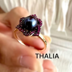 May include: A gold ring featuring a dark blue pearl surrounded by small, deep red gemstones. The ring's setting is shaped like a flower. The word "THALIA" is printed in black letters at the bottom of the image.