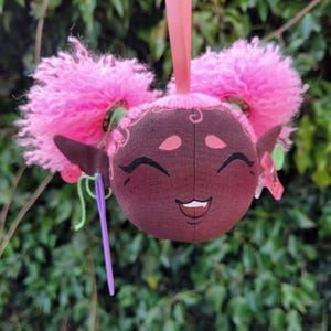 May include: A plush toy with pink hair, brown skin, and a smiling face. The toy is wearing a green and red beaded necklace and has a purple needle and green yarn attached to its ear.