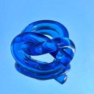 Puede incluir: Una escultura de vidrio azul en forma de nudo. La escultura está hecha de vidrio transparente con un tinte azul. Está sentada sobre una superficie azul.