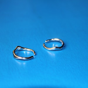 May include: Two silver rings with an open design and a flowing, organic shape. They sit on a bright blue surface, which accentuates their metallic shine. The rings have a minimalist, modern aesthetic. The jewellery is likely for the ear.