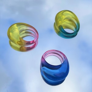 Op de afbeelding: Drie kleurrijke ringen gemaakt van gelaagd plastic. De ringen zijn in tinten geel, blauw en roze.