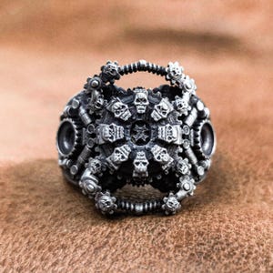 May include: A silver ring with a detailed design of skulls and gears. The ring is made of metal and has a steampunk aesthetic.