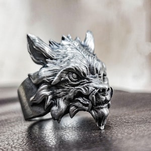 Puede incluir: Anillo plateado con un diseño detallado de cabeza de lobo. El anillo muestra la cara del lobo con texturas intrincadas, incluyendo pelaje y dientes afilados. Una pieza audaz para quienes aprecian la joyería con temática animal.