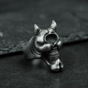 Puede incluir: Un anillo de plata con un diseño de calavera detallado con cuernos. La calavera tiene una amplia sonrisa con dientes y una gran cuenca del ojo vacía.