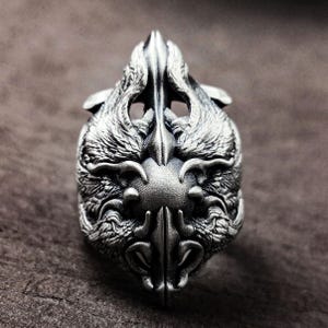 Op de afbeelding: Een zilveren ring met een gedetailleerd ontwerp van twee gevederde wezens die elkaar aankijken. De ring is gemaakt van metaal en heeft een getextureerd oppervlak.