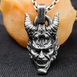 Puede incluir: Un collar con un colgante de plata que presenta una escultura detallada en tres dimensiones de la cabeza de un demonio con cuernos.