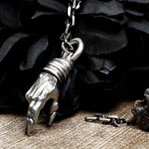 Könnte beinhalten: Ein silberner handförmiger Anhänger hängt an einer silbernen Kette. Der Anhänger ist mit strukturierten Linien und einem spitzen Finger verziert. Die Kette ist mit einer größeren silbernen Kette verbunden.