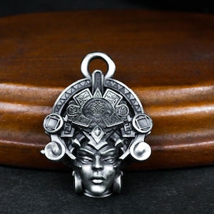 Puede incluir: Un colgante de plata con un rostro estilizado detallado con intrincados diseños geométricos a su alrededor. El colgante tiene un bucle en la parte superior para sujetarlo a una cadena.