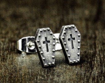 May include: A pair of silver coffin-shaped earrings with a cross design. The earrings have a textured surface and are studded with small silver dots.