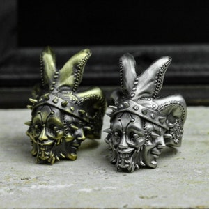 May include: Two silver rings with a three-faced jester design. The jester has a pointed hat, a stitched face, and a toothy grin. One ring is gold-toned and the other is silver.