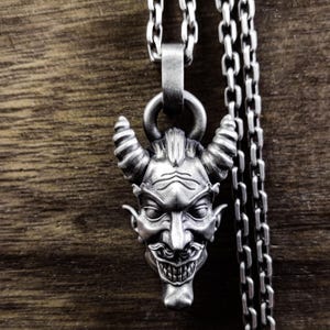 May include: A silver pendant necklace featuring a detailed, three-dimensional sculpture of a devil's face with horns. The pendant is attached to a silver chain.