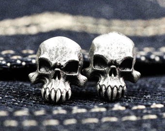 Può includere: Un paio di orecchini a forma di teschio in argento con dettagli elaborati, inclusi denti e orbite. Gli orecchini sono mostrati su uno sfondo in tessuto denim scuro.