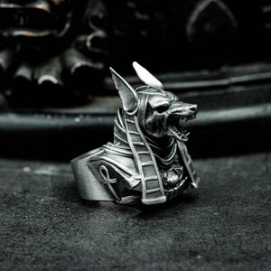 K&ouml;nnte beinhalten: Ein silberfarbener Ring mit dem Kopf von Anubis, dem &auml;gyptischen Gott. Der Ring zeigt Anubis mit detaillierten Merkmalen, einschlie&szlig;lich spitzer Ohren und offenem Mund. Der Ring hat ein detailliertes Hieroglyphen-Design auf der Brust.