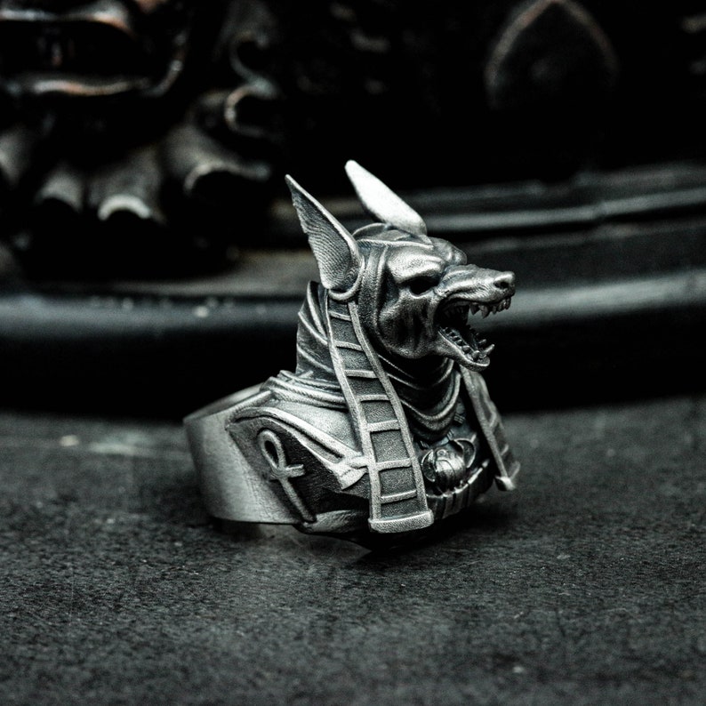 K&ouml;nnte beinhalten: Ein silberfarbener Ring mit dem Kopf von Anubis, dem &auml;gyptischen Gott. Der Ring zeigt Anubis mit detaillierten Merkmalen, einschlie&szlig;lich spitzer Ohren und offenem Mund. Der Ring hat ein detailliertes Hieroglyphen-Design auf der Brust.