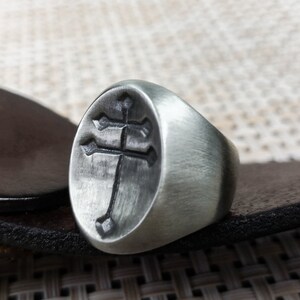 May include: A silver ring featuring a cross design. The ring is oval shaped and has a brushed finish.