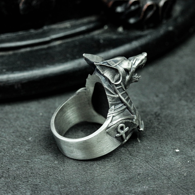 K&ouml;nnte beinhalten: Ein silberner Ring mit detailliertem Wolfskopf-Design. Der Ring hat eine offene R&uuml;ckseite und ein Ankh-Symbol an der Seite. Das Metall hat eine geb&uuml;rstete Oberfl&auml;che, die ihm ein Vintage-Aussehen verleiht. Ein auff&auml;lliges Schmuckst&uuml;ck.