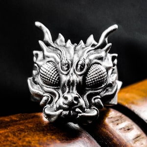 May include: A silver ring featuring a detailed dragon head design. The dragon has large, round eyes and a fierce expression.