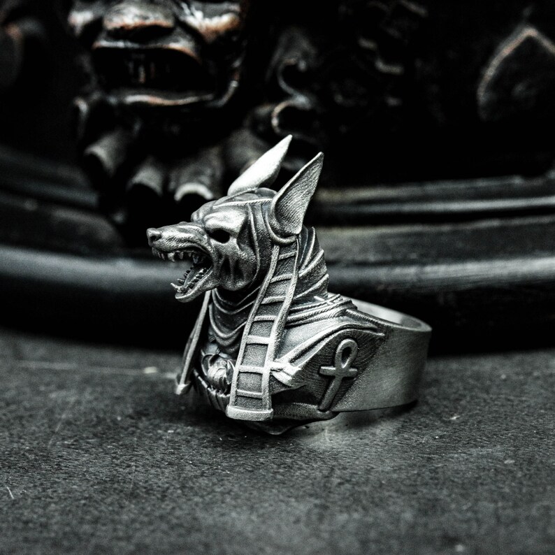 K&ouml;nnte beinhalten: Ein silberner Ring mit dem Kopf von Anubis, dem &auml;gyptischen Gott, mit grimmigem Gesichtsausdruck und detaillierten Hieroglyphen. Der Ring hat eine polierte Oberfl&auml;che und ein Ankh-Symbol auf dem Band.