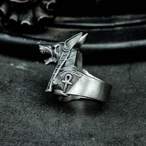 K&ouml;nnte beinhalten: Silberfarbener Ring mit dem Kopf von Anubis, dem &auml;gyptischen Gott der Toten, mit detaillierten Gesichtsz&uuml;gen und einem Ankh-Symbol. Der Ring hat ein breites Band und eine strukturierte Oberfl&auml;che, die ein gotisches oder alt&auml;gyptisches Thema aufweist.