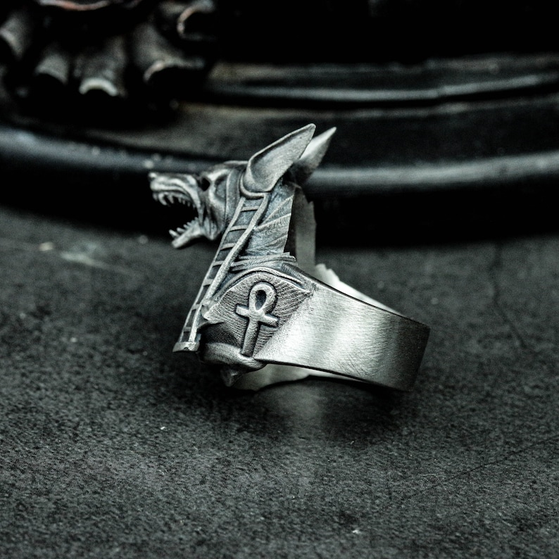 K&ouml;nnte beinhalten: Silberfarbener Ring mit dem Kopf von Anubis, dem &auml;gyptischen Gott der Toten, mit detaillierten Gesichtsz&uuml;gen und einem Ankh-Symbol. Der Ring hat ein breites Band und eine strukturierte Oberfl&auml;che, die ein gotisches oder alt&auml;gyptisches Thema aufweist.