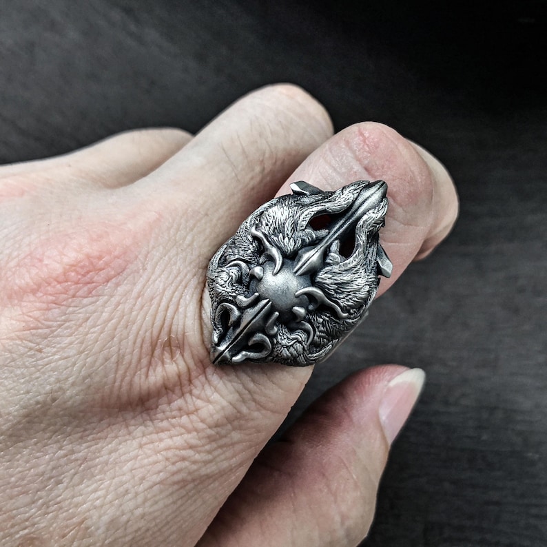 May include: A silver ring with a detailed design of two wolf heads facing each other. The ring is worn on a finger.