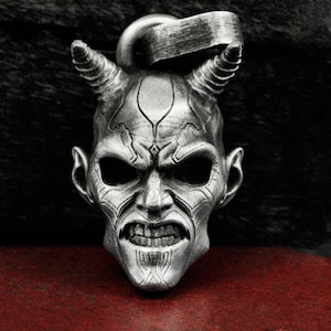 Może przedstawiać: Srebrny wisiorek w kształcie głowy demona z rogami. Demon ma groźny wyraz twarzy z szeroko otwartymi ustami i ostrymi zębami.