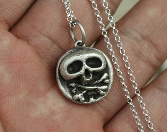 May include: Silver skull and crossbones pendant necklace. The pendant is round and features a detailed skull with crossed bones. The necklace is made of a silver chain.