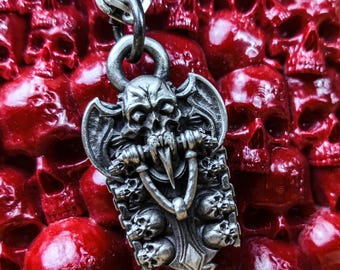 May include: A silver pendant necklace featuring a detailed skull with wings and a ring in its mouth. The pendant is surrounded by smaller skulls and is hanging against a background of red skulls.