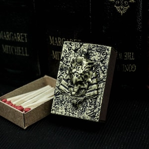Puede incluir: Una caja de cerillas de color dorado con un diseño en relieve de una criatura mítica con una corona y un fondo detallado. La caja de cerillas está cerrada y se encuentra sobre una superficie oscura.
