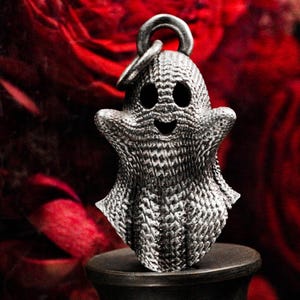 Puede incluir: Un amuleto de plata con forma de fantasma sonriente con una superficie texturizada. El amuleto tiene un bucle en la parte superior para sujetarlo a un collar o pulsera.