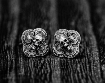 May include: A pair of silver skull earrings with a clover-shaped design. The earrings are detailed with intricate patterns and feature a skull in the center of each clover.