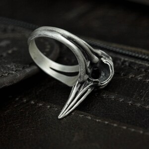 Könnte beinhalten: Ein silberner Ring mit einem detaillierten Schädel-Design. Der Schädel hat einen spitzen Schnabel und komplizierte Details. Der Ring liegt auf einer dunkelbraunen Lederoberfläche.