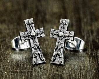 May include: A pair of silver-toned cross stud earrings. Each earring features an ornate cross design with floral and fleur-de-lis details. The earrings are set against a dark, textured background.