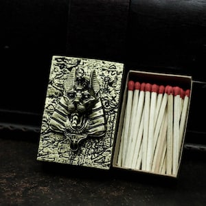 Puede incluir: Una caja de cerillas de estilo vintage con una tapa de metal dorado con una imagen en relieve de un gato egipcio con un tocado de faraón. La caja de cerillas está abierta, revelando una caja llena de cerillas con puntas rojas.