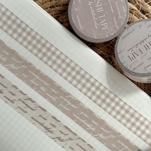 May include: A notebook with grid paper displays strips of beige and brown washi tape, including a gingham pattern and script text. Several rolls of washi tape are visible, with the text "WASHI TAPE by artstudiolea". The tape is ideal for crafts and journaling.