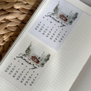 Könnte beinhalten: Zwei transparente Aufkleber mit einem Januar-Kalenderdesign. Jeder Aufkleber zeigt eine Aquarellillustration einer Hütte in einem verschneiten Wald. Das Wort "Januar" ist in eleganter Schrift über dem Kalenderraster geschrieben.