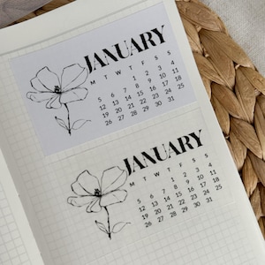 May include: Two transparent stickers with the month of January printed in black, each featuring a floral illustration. The stickers are placed on a grid-lined notebook page. The calendar includes the days of the month.
