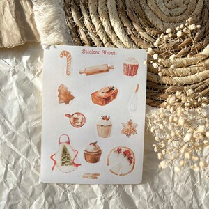 Könnte beinhalten: Eine Sticker-Tafel mit verschiedenen Illustrationen zum Thema Weihnachtsbacken. Die Tafel enthält einen Nudelholz, ein Lebkuchenhaus, einen Zuckerstange, einen Cupcake, ein Brot, einen Kuchen, einen Schneebesen, einen Lebkuchen und eine Schürze mit einem Weihnachtsbaum-Motiv. Die Tafel ist weiß mit rotem Text, der "Sticker Sheet" sagt.