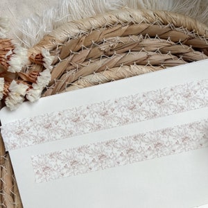 Könnte beinhalten: Zwei Rollen weißes Washi-Tape mit einem zarten Blumenmuster in brauner Tinte. Das Band befindet sich auf einer weißen Oberfläche.