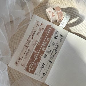Könnte beinhalten: Eine Sammlung dekorativer Washi-Tapes im Vintage-Stil. Die Tapes zeigen braune und weiße Designs mit Zahlen, Text und Illustrationen. Die Tapes befinden sich auf einem weißen Notizbuch mit Rastermuster.