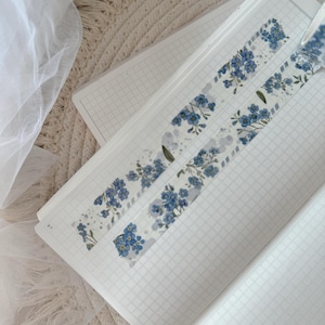 Könnte beinhalten: Zwei Rollen blaues und weißes Washi-Tape mit einem floralen Muster aus Vergissmeinnicht. Das Band befindet sich auf einem weißen karierten Notizbuch.
