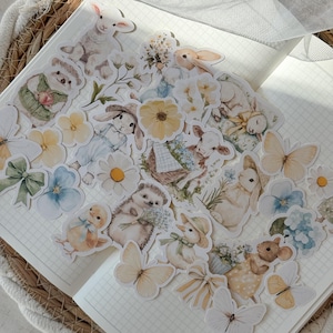 Könnte beinhalten: Eine Sammlung pastellfarbener Aufkleber mit verschiedenen Frühlingsillustrationen. Die Aufkleber zeigen Hasen, Lämmer, Schmetterlinge, Blumen und andere bezaubernde Designs. Die Aufkleber sind auf einer Notizbuchseite angeordnet und schaffen eine bezaubernde Szene.