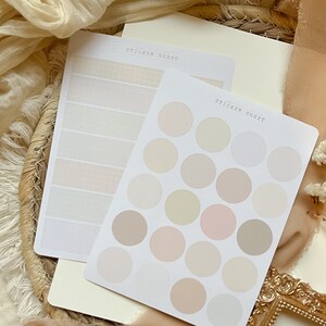 Könnte beinhalten: Zwei Stickerbögen mit einer neutralen Farbpalette. Ein Bogen hat 18 runde Sticker in verschiedenen Beige-, Creme- und Rosatönen. Der andere Bogen hat 6 rechteckige Sticker mit einer dezenten Leinenstruktur.