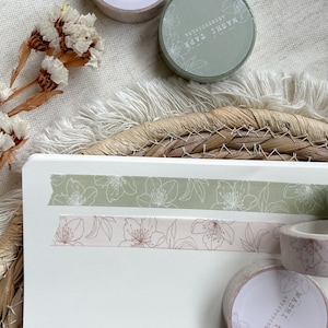 Könnte beinhalten: Zwei Rollen Washi-Tape mit Blumenmustern. Die obere Rolle ist grün mit weißen Blumen und die untere Rolle ist rosa mit braunen Blumen. Der Text auf den Rollen lautet "Washi Tape" und "Botanical".