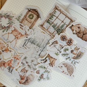 Könnte beinhalten: Eine Sammlung von Aufklebern zum Thema Winter mit Waldtieren und verschneiten Szenen. Die Aufkleber zeigen ein Reh, einen Fuchs, ein Eichhörnchen, einen Bären und einen Igel sowie eine Hütte, ein Fenster und Tannenzapfen. Die Farbpalette ist sanft, mit gedämpften Brauntönen, Weiß und Grün.