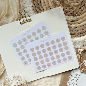 Könnte beinhalten: Zwei Blätter beige und weißer Aufkleber mit dem Wort "Essentials" obenauf gedruckt. Jedes Blatt hat 31 beige Kreise mit den Zahlen 1 bis 31, die in Schwarz in jeden Kreis gedruckt sind.